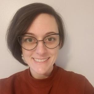 A white woman with chin-length, straight brown hair, horn-rimmed glasses, and a terracotta colored sweater smiles into the camera.