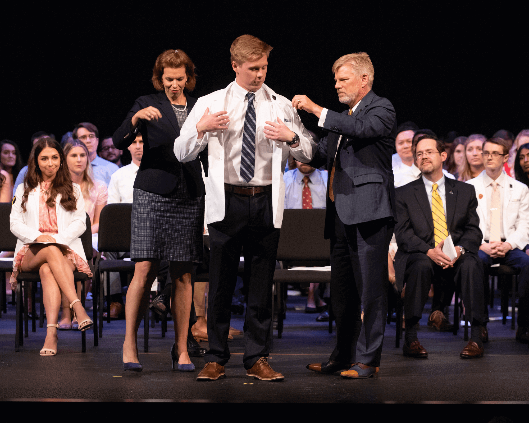 Faculty helping a medical student put on his white coat.