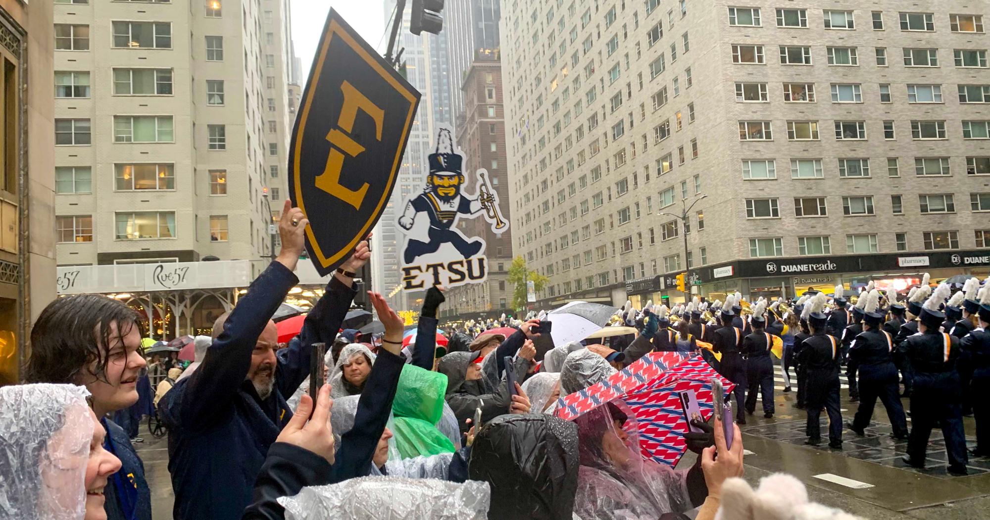 Spectators in ponchos cheer and hold ETSU signs as the Marching Bucs pass by during the Macy's Thanksgiving Day Parade in a rainy New York City.