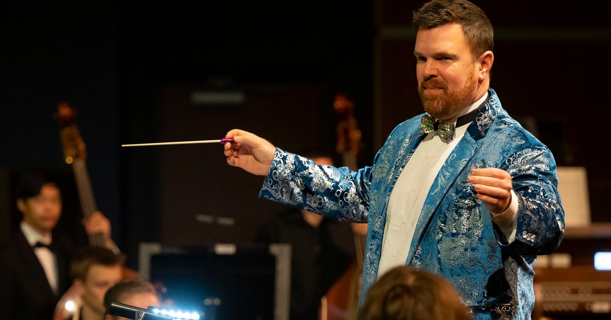 Dr. Alan Stevens conducts an orchestra with a baton, wearing a vibrant blue paisley jacket and bow tie during a performance.