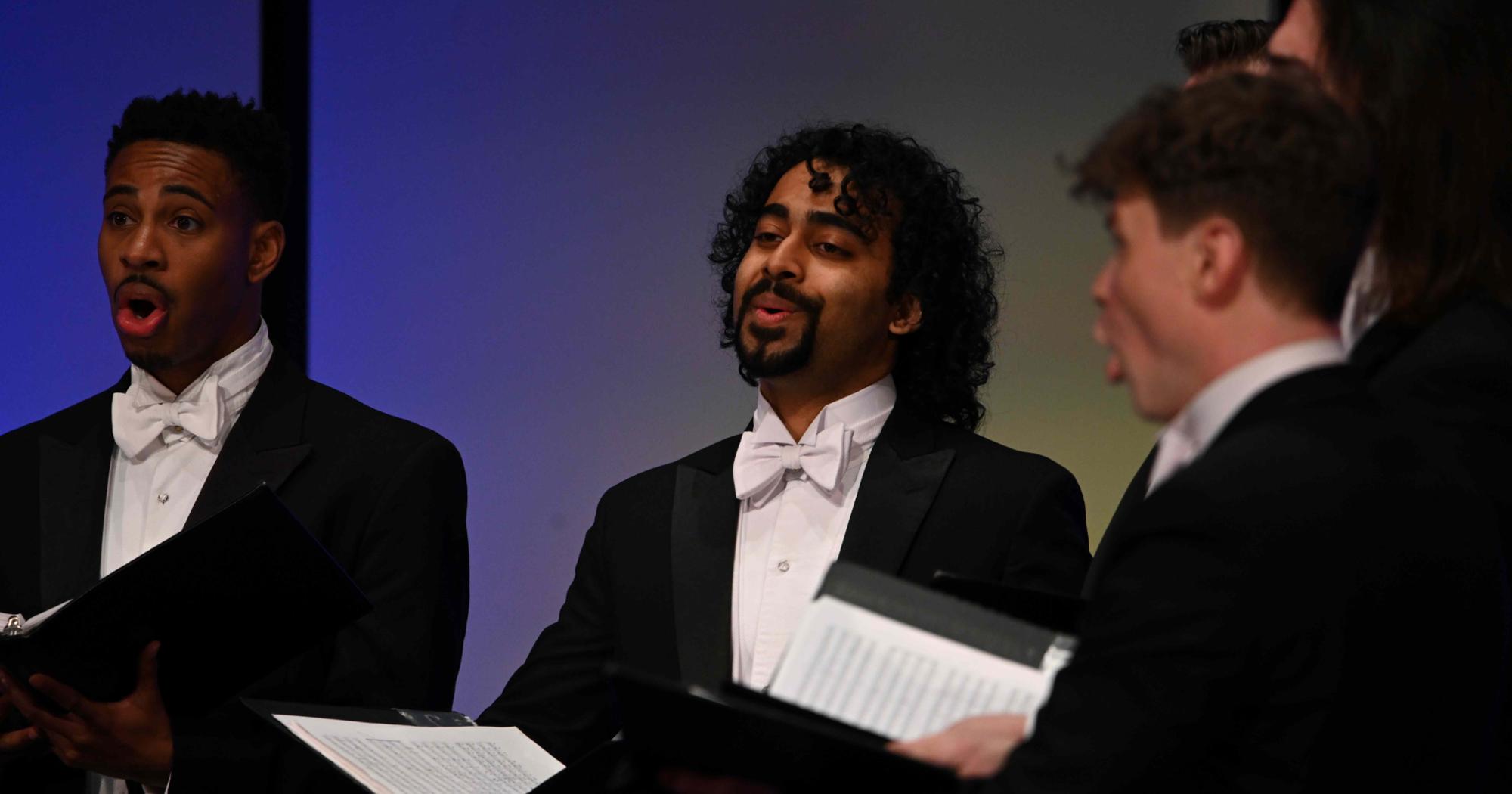 Chanticleer vocalists in formal attire perform on stage, passionately singing with open music folders during their 2023 concert.