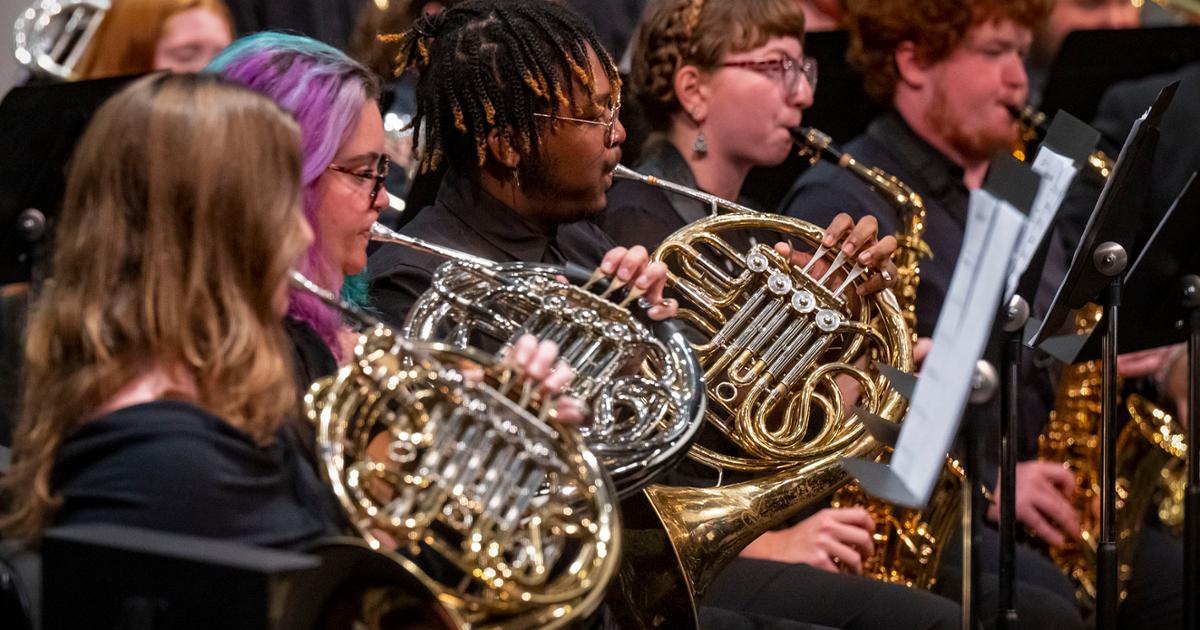 Musicians play French horns in unison during a closing band concert, seated alongside other brass instrument performers.